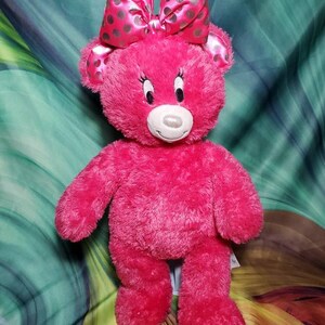 May include: A plush pink teddy bear with a large bow featuring silver polka dots. The bear has a white muzzle, black eyes, and a friendly expression. This soft toy is a charming gift for children.
