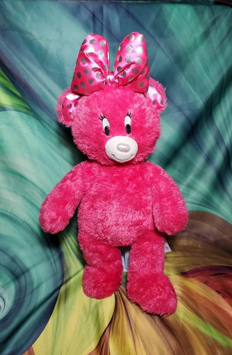 May include: A plush pink teddy bear with a large bow featuring silver polka dots. The bear has a white muzzle, black eyes, and a friendly expression. This soft toy is a charming gift for children.