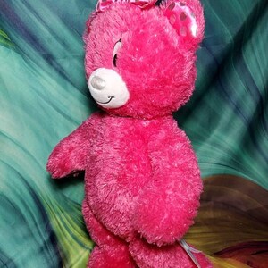 May include: A bright pink plush teddy bear with a matching bow featuring black polka dots. The bear has a white muzzle and a smiling expression. This stuffed animal is a cute gift for children.
