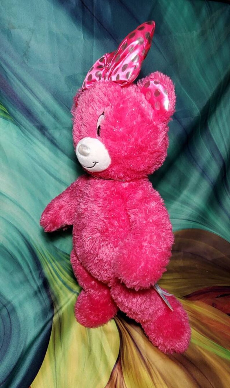May include: A bright pink plush teddy bear with a matching bow featuring black polka dots. The bear has a white muzzle and a smiling expression. This stuffed animal is a cute gift for children.