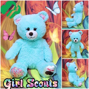 May include: A large, light blue teddy bear with a white muzzle, black eyes, and patterned ears and foot pads. The bear has the words "Thin Mints" on one foot and the words "Girl Scouts" at the bottom of the image. Butterflies are in the background.