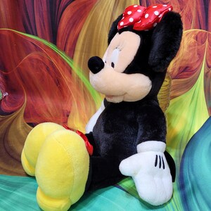 K&ouml;nnte beinhalten: Eine Pl&uuml;schpuppe von Minnie Mouse mit schwarzem Fell, einem wei&szlig;en Gesicht und gro&szlig;en gelben F&uuml;&szlig;en. Sie tr&auml;gt eine rote Schleife mit wei&szlig;en Punkten und wei&szlig;e Handschuhe. Ein klassischer Disney-Charakter.