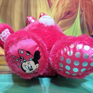 Puede incluir: Un peluche rosa brillante con Minnie Mouse. El juguete tiene una textura suave y difusa con detalles de sat&eacute;n. Minnie Mouse aparece en las almohadillas de los pies, que est&aacute;n decoradas con lunares. Tambi&eacute;n son visibles las orejas y el lazo del juguete.