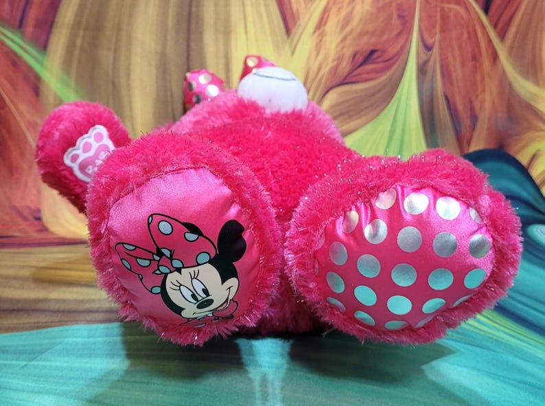 Puede incluir: Un peluche rosa brillante con Minnie Mouse. El juguete tiene una textura suave y difusa con detalles de sat&eacute;n. Minnie Mouse aparece en las almohadillas de los pies, que est&aacute;n decoradas con lunares. Tambi&eacute;n son visibles las orejas y el lazo del juguete.