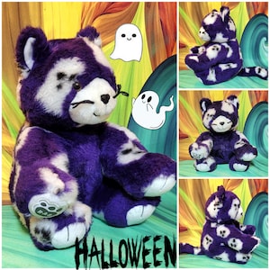 May include: A plush cat toy featuring purple and white fur with a repeating ghost pattern. The cat has black embroidered whiskers, a black nose, and a white paw pad with a B-A-B logo. The word HALLOWEEN is printed in black at the bottom.