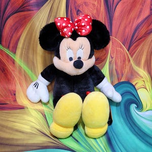 K&ouml;nnte beinhalten: Eine Pl&uuml;schpuppe von Minnie Mouse, mit einem schwarzen K&ouml;rper, gro&szlig;en schwarzen Ohren und einer roten Schleife mit wei&szlig;en Punkten. Die Puppe hat wei&szlig;e Handschuhe und gelbe Schuhe. Der Hintergrund ist ein farbenfrohes abstraktes Design.