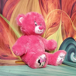 K&ouml;nnte beinhalten: Ein pinkfarbener Teddyb&auml;r mit wei&szlig;er Schnauze und Innenohren. Die Pfoten des B&auml;ren zeigen ein Minnie Mouse-Design mit einer rosa Schleife. Der Hintergrund hat ein farbenfrohes, abstraktes Blumenmuster.