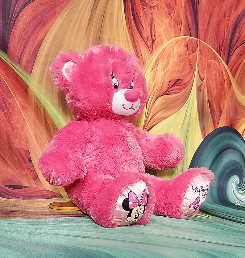 K&ouml;nnte beinhalten: Ein pinkfarbener Teddyb&auml;r mit wei&szlig;er Schnauze und Innenohren. Die Pfoten des B&auml;ren zeigen ein Minnie Mouse-Design mit einer rosa Schleife. Der Hintergrund hat ein farbenfrohes, abstraktes Blumenmuster.
