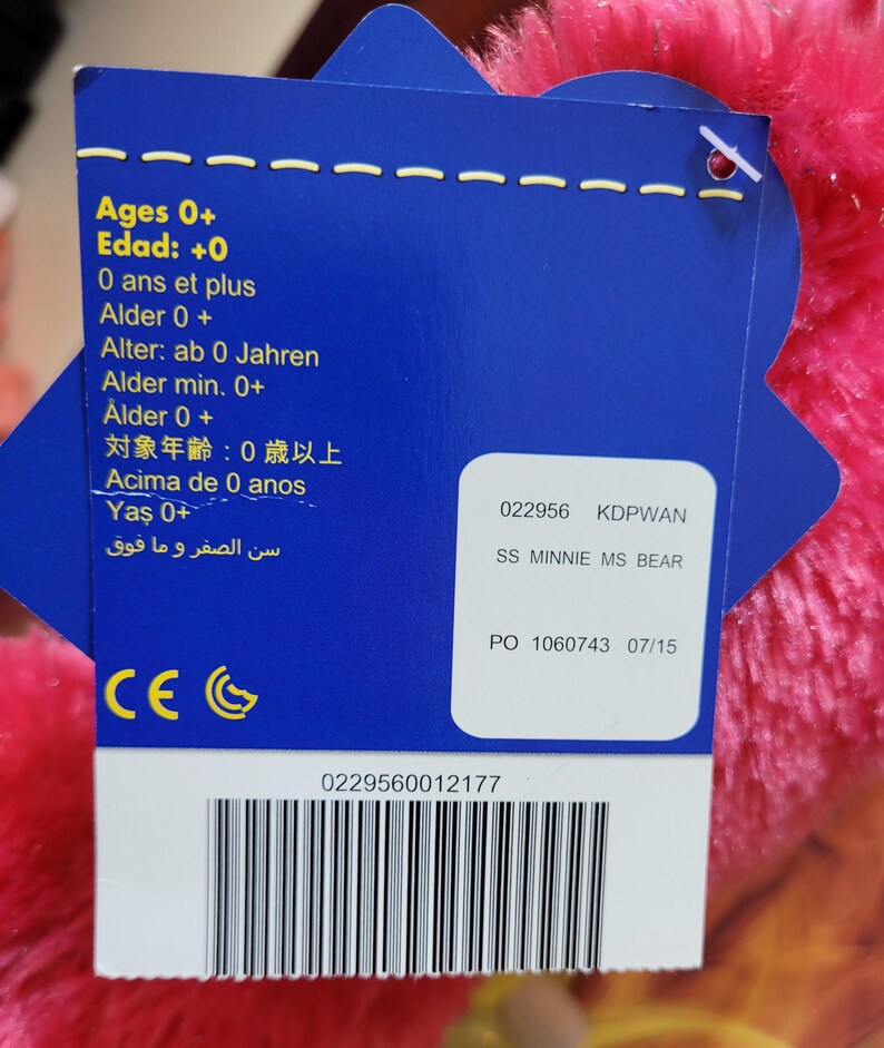 Puede incluir: Una etiqueta azul con texto en varios idiomas, incluyendo "Ages 0+" y "SS MINNIE MS BEAR." La etiqueta est&aacute; adjunta a un peluche rosa y esponjoso. La etiqueta tambi&eacute;n incluye un c&oacute;digo de barras y el n&uacute;mero "0229560012177."