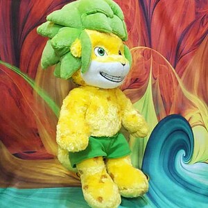 May include: A plush toy lion with a yellow body, green hair, and green shorts. The lion has a smiling face with white fur and green eyes. The background features a colorful, abstract design with red, orange, and blue hues.