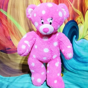 May include: Pink plush teddy bear with white polka dots. The bear has a pink nose and black eyes with eyelashes.