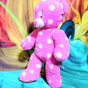May include: Pink plush teddy bear with white polka dots. The bear is standing on a blue surface.