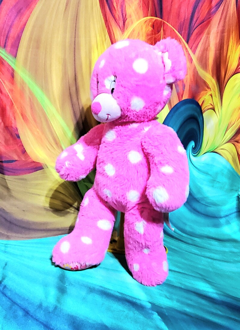 May include: Pink plush teddy bear with white polka dots. The bear is standing on a blue surface.