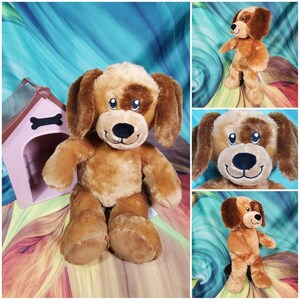 May include: A brown and tan plush dog toy with a brown nose and black eyes. The dog is sitting down with its paws in front of it. The dog is in front of a pink and brown dog house.