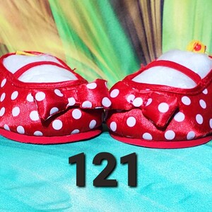 K&ouml;nnte beinhalten: Ein Paar rote Babyschuhe mit wei&szlig;en Punkten und einer Schleife auf der Vorderseite. Die Schuhe haben eine wei&szlig;e Innenseite und einen roten Riemen. Die Nummer "121" wird in Schwarz am unteren Bildrand angezeigt.