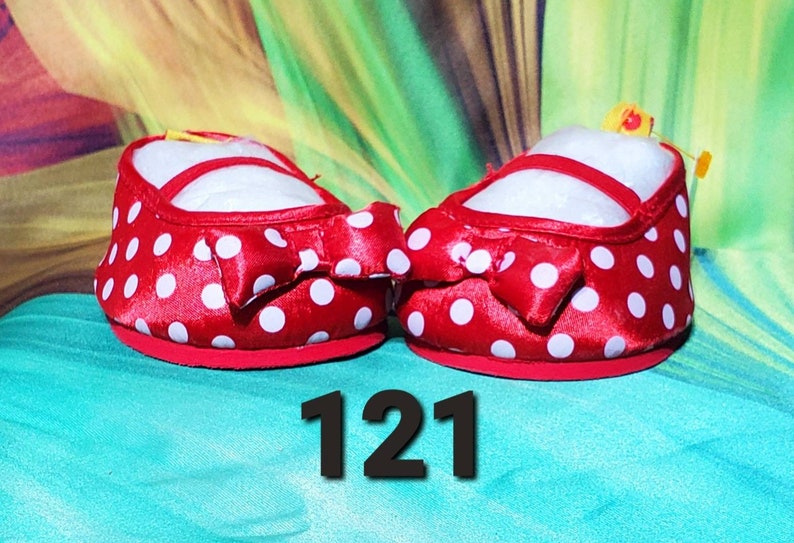 K&ouml;nnte beinhalten: Ein Paar rote Babyschuhe mit wei&szlig;en Punkten und einer Schleife auf der Vorderseite. Die Schuhe haben eine wei&szlig;e Innenseite und einen roten Riemen. Die Nummer "121" wird in Schwarz am unteren Bildrand angezeigt.