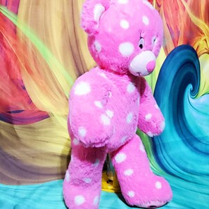 May include: A pink plush teddy bear with white polka dots. The bear is standing on a colorful background.