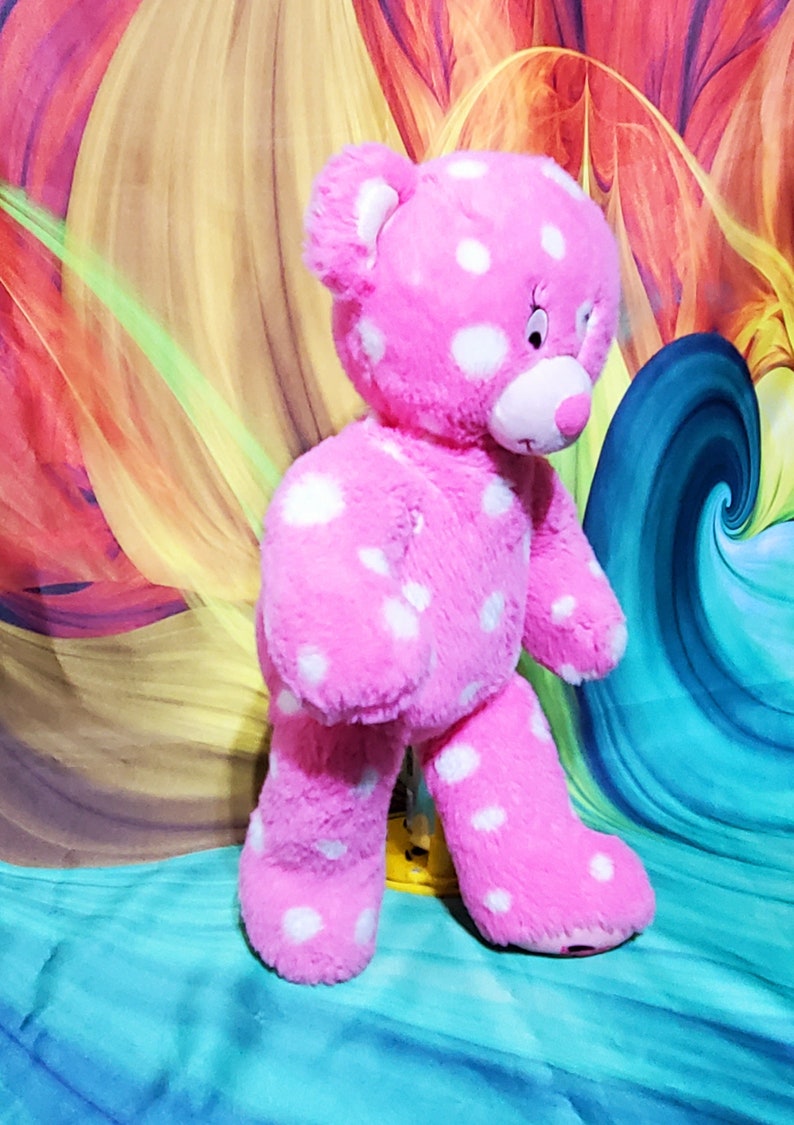 May include: A pink plush teddy bear with white polka dots. The bear is standing on a colorful background.