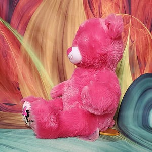 K&ouml;nnte beinhalten: Ein rosa Pl&uuml;schteddyb&auml;r mit wei&szlig;er Schnauze und l&auml;chelndem Gesicht. Der B&auml;r hat ein Minnie-Maus-Design auf seinem Fu&szlig;. Der B&auml;r sitzt auf einer blau-gr&uuml;nen Oberfl&auml;che mit farbenfrohem Hintergrund.