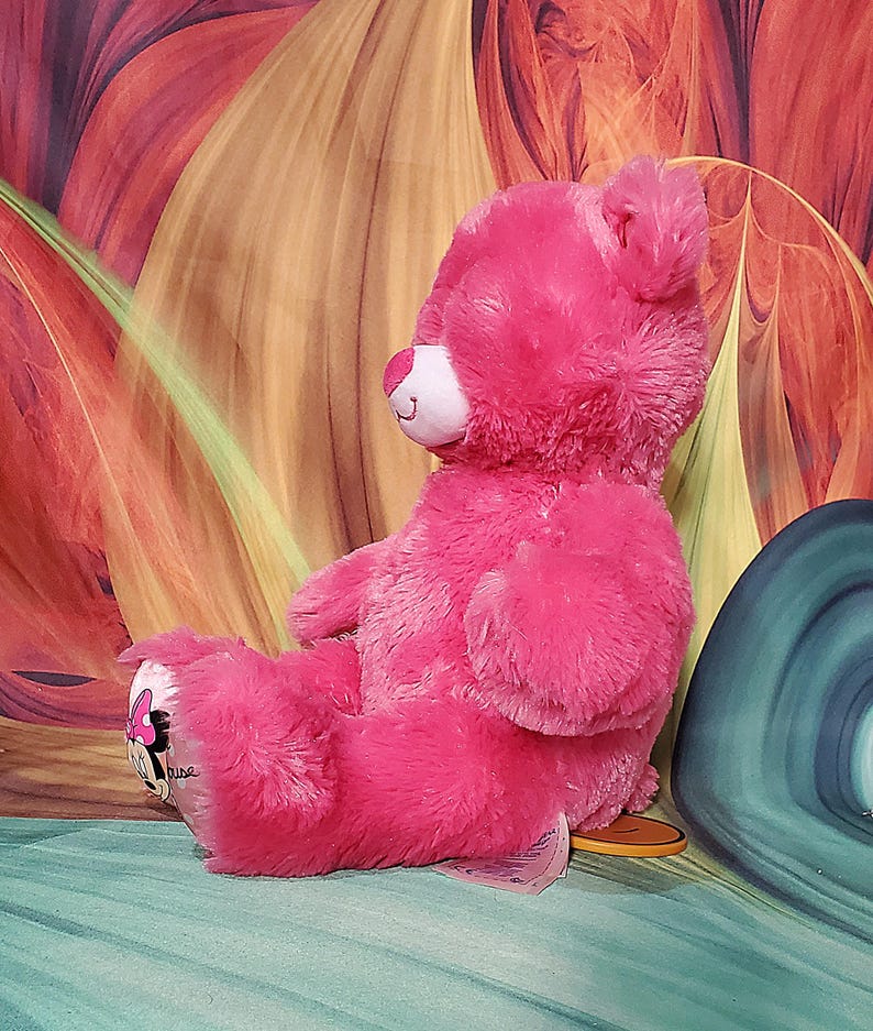 K&ouml;nnte beinhalten: Ein rosa Pl&uuml;schteddyb&auml;r mit wei&szlig;er Schnauze und l&auml;chelndem Gesicht. Der B&auml;r hat ein Minnie-Maus-Design auf seinem Fu&szlig;. Der B&auml;r sitzt auf einer blau-gr&uuml;nen Oberfl&auml;che mit farbenfrohem Hintergrund.