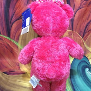 Puede incluir: Un oso de peluche rosa brillante con un lazo a juego con lunares dorados. El oso tiene una textura suave y esponjosa y extremidades peque&ntilde;as. Una etiqueta est&aacute; adjunta al oso.
