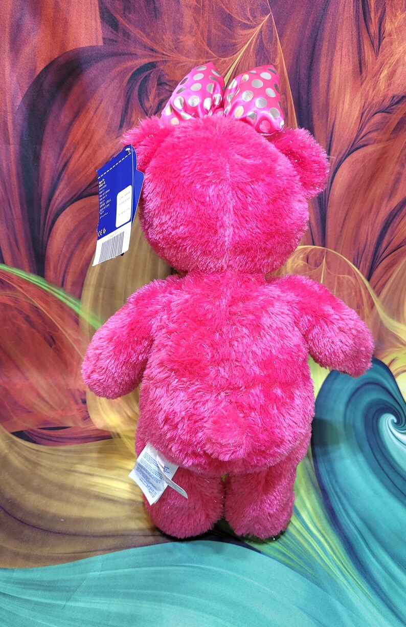 Puede incluir: Un oso de peluche rosa brillante con un lazo a juego con lunares dorados. El oso tiene una textura suave y esponjosa y extremidades peque&ntilde;as. Una etiqueta est&aacute; adjunta al oso.