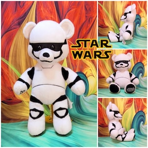 May include: A white plush bear featuring black accents designed to resemble a Star Wars Stormtrooper armor suit. The bear has a black mask detail around the eyes, black limb markings, and the Star Wars logo is printed on the right foot pad.