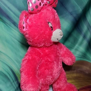 May include: A bright pink plush teddy bear with a large bow featuring silver polka dots. The bear has a white muzzle and a smiling expression. The toy is designed for children.