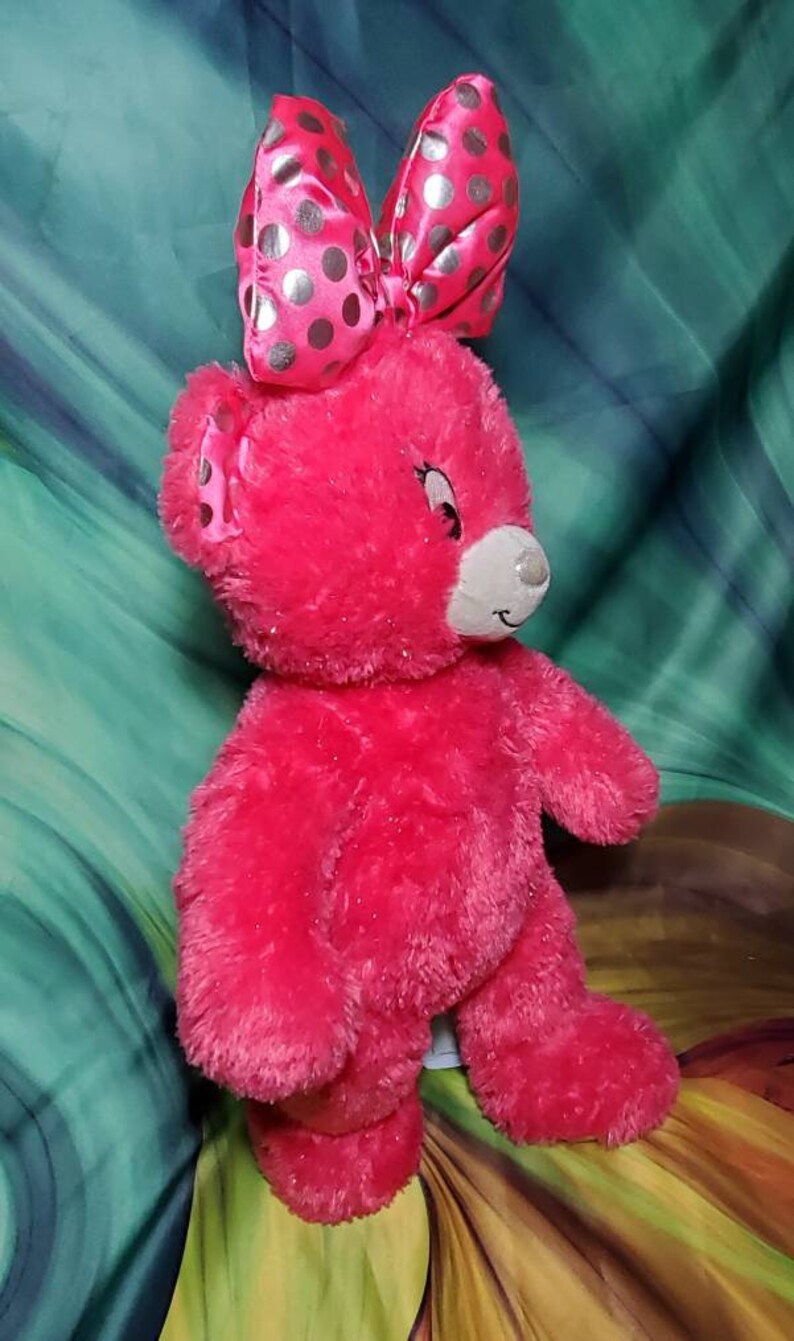 May include: A bright pink plush teddy bear with a large bow featuring silver polka dots. The bear has a white muzzle and a smiling expression. The toy is designed for children.