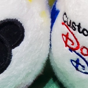 Pu&ograve; includere: Due palline di peluche bianche con accenti colorati. Una pallina presenta una silhouette nera di Topolino. L'altra pallina ha il testo ricamato "Custom made at DOWNTOWN DISNEY" in rosso, blu e nero.