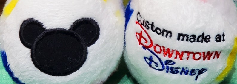 Pu&ograve; includere: Due palline di peluche bianche con accenti colorati. Una pallina presenta una silhouette nera di Topolino. L'altra pallina ha il testo ricamato "Custom made at DOWNTOWN DISNEY" in rosso, blu e nero.
