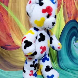 Pu&ograve; includere: Un orsacchiotto di peluche bianco con forme astratte colorate in rosso, giallo, blu e nero. L'orso ha un'espressione amichevole ed &egrave; in piedi. Lo sfondo presenta un design astratto vibrante.