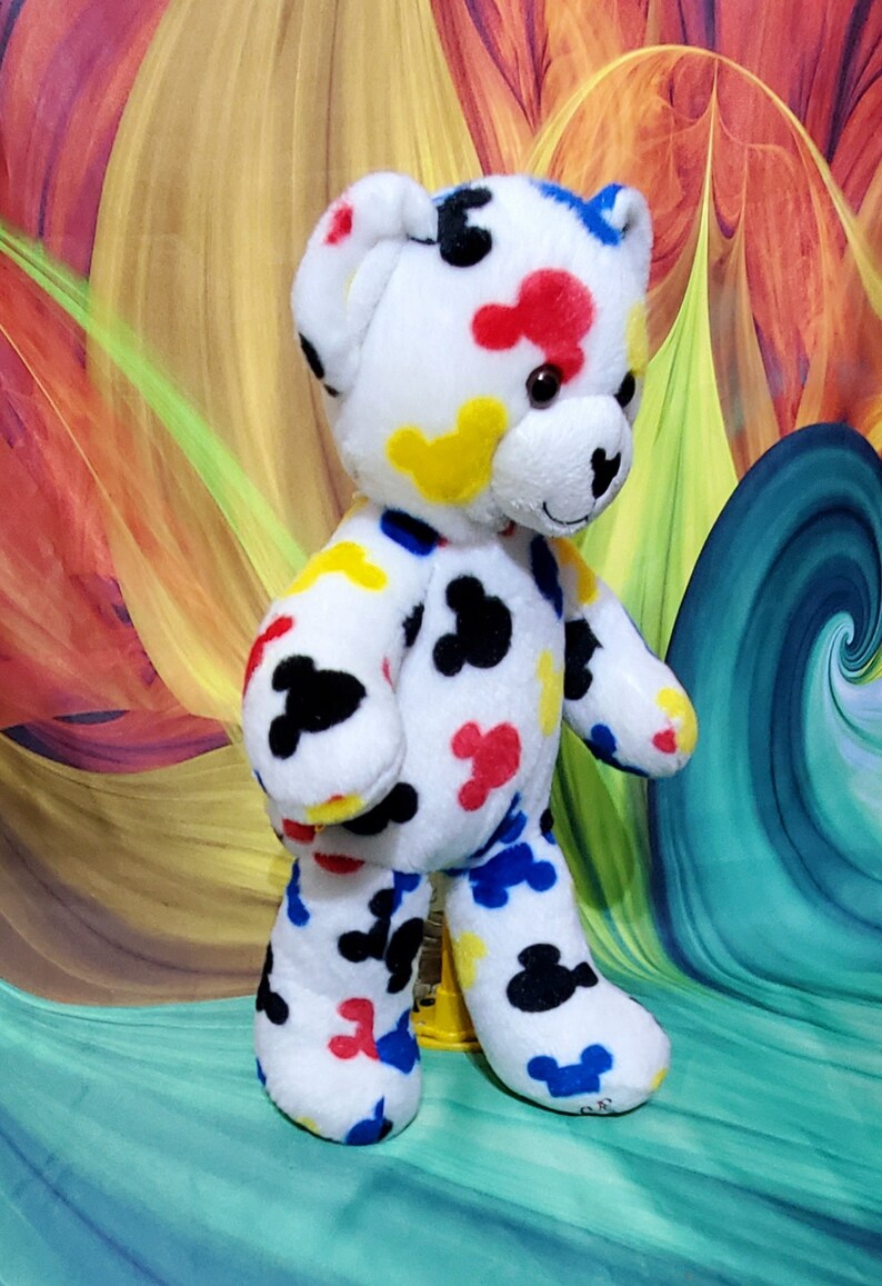 Pu&ograve; includere: Un orsacchiotto di peluche bianco con forme astratte colorate in rosso, giallo, blu e nero. L'orso ha un'espressione amichevole ed &egrave; in piedi. Lo sfondo presenta un design astratto vibrante.