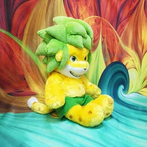May include: A plush toy lion with a yellow body, green shorts, and a green leafy mane. The lion has a smiling face with yellow eyes and is sitting on a turquoise surface. The background features a colorful abstract design with red, yellow, and orange hues.