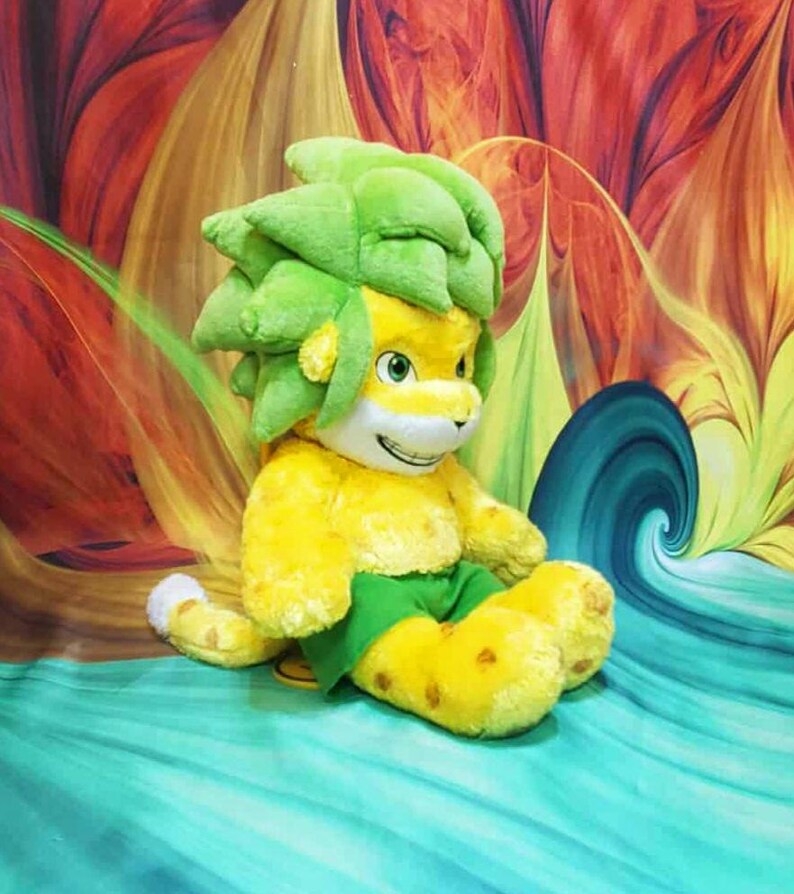 May include: A plush toy lion with a yellow body, green shorts, and a green leafy mane. The lion has a smiling face with yellow eyes and is sitting on a turquoise surface. The background features a colorful abstract design with red, yellow, and orange hues.