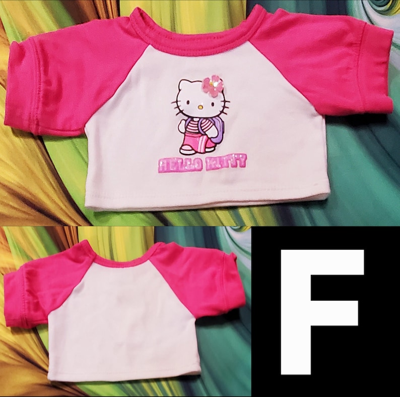 Może przedstawiać: Biała i r&oacute;żowa koszulka w stylu raglan z grafiką Hello Kitty. Koszulka ma r&oacute;żowy dekolt i rękawy, z postacią Hello Kitty i napisem "Hello Kitty" wydrukowanym z przodu.