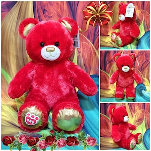 May include: A plush red teddy bear with gold accents on the ears and paws. The bear has a white muzzle and a gold nose. The bear's paws have the letters "B-A-B" printed on them. The bear is surrounded by a colorful background.