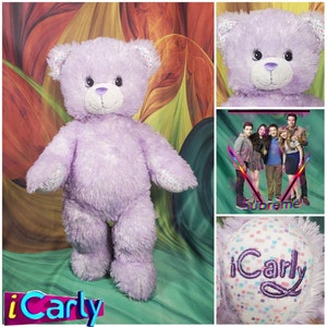 May include: Purple plush iCarly bear with a white face and sparkly details. The bear is standing on a green and orange background. The iCarly logo is visible in the bottom left corner of the image.