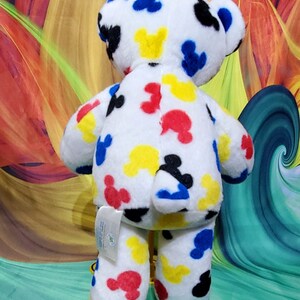 Pu&ograve; includere: Un orsacchiotto di peluche bianco con motivi colorati di Mickey Mouse in rosso, giallo, blu e nero. L'orso ha un'etichetta attaccata alla zampa. Lo sfondo presenta un design astratto colorato.
