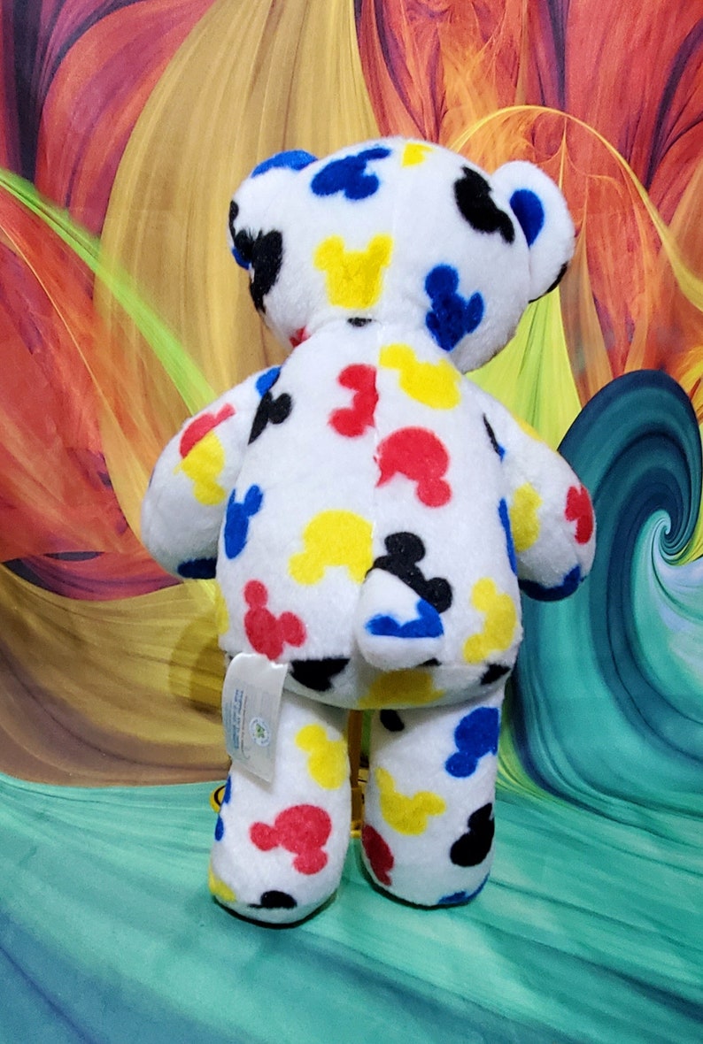 Pu&ograve; includere: Un orsacchiotto di peluche bianco con motivi colorati di Mickey Mouse in rosso, giallo, blu e nero. L'orso ha un'etichetta attaccata alla zampa. Lo sfondo presenta un design astratto colorato.