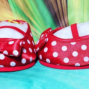 K&ouml;nnte beinhalten: Ein Paar rote Babyschuhe mit wei&szlig;en Punkten. Jeder Schuh hat eine rote Schleife und einen Riemen. Die Schuhe stehen vor einem gr&uuml;n-gelben Hintergrund. Ein Etikett mit der Nummer "121" ist an einem Schuh befestigt.