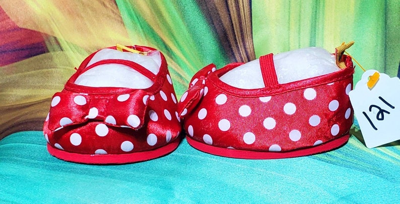 K&ouml;nnte beinhalten: Ein Paar rote Babyschuhe mit wei&szlig;en Punkten. Jeder Schuh hat eine rote Schleife und einen Riemen. Die Schuhe stehen vor einem gr&uuml;n-gelben Hintergrund. Ein Etikett mit der Nummer "121" ist an einem Schuh befestigt.