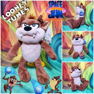 May include: A plush Taz, the Tasmanian Devil, from Looney Tunes. The brown and tan plush toy has a stern expression, with a tag attached. The image also includes the Space Jam: A New Legacy logo and cartoon illustrations of Taz.