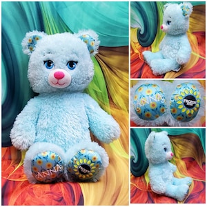 May include: A light blue plush bear with blue eyes, a pink nose, and a smiling mouth. The bear's ears and foot pads feature a sunflower pattern. The foot pads also display the text "ANNA" and "FROZEN FEVER".