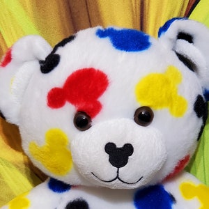 Pu&ograve; includere: Un orsacchiotto di peluche bianco con macchie colorate a forma di Mickey Mouse in rosso, blu, giallo e nero. L'orso ha occhi marroni e un naso nero. Le orecchie e il viso dell'orso sono dettagliati.