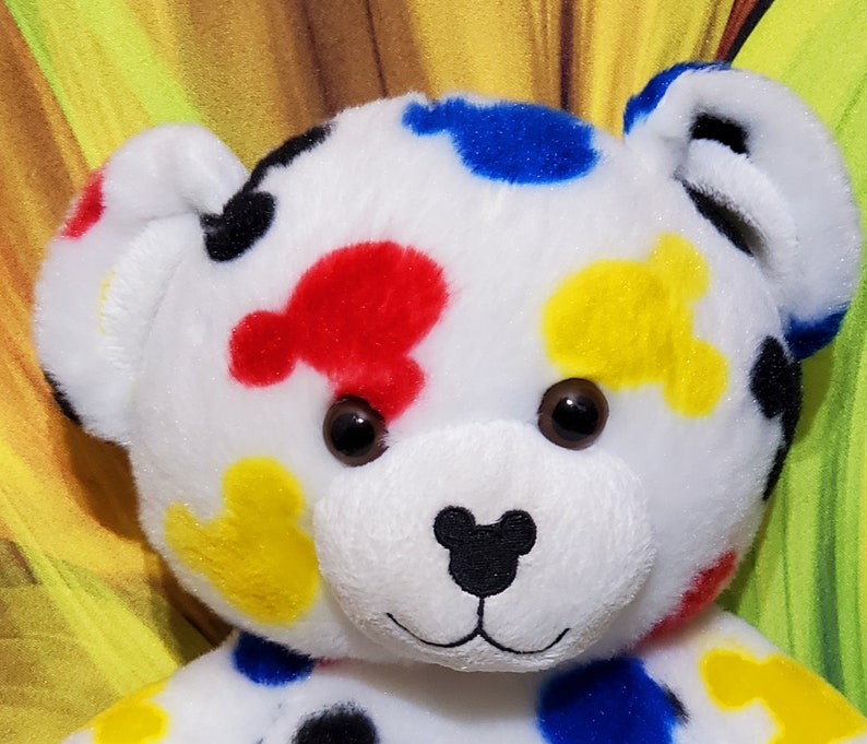 Pu&ograve; includere: Un orsacchiotto di peluche bianco con macchie colorate a forma di Mickey Mouse in rosso, blu, giallo e nero. L'orso ha occhi marroni e un naso nero. Le orecchie e il viso dell'orso sono dettagliati.