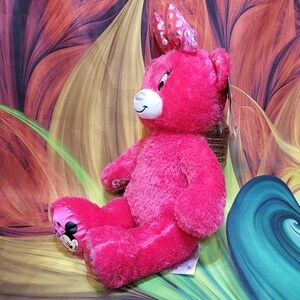 Puede incluir: Un oso de peluche rosa brillante con un lazo en la cabeza y un gr&aacute;fico de Minnie Mouse en el pie. El oso tiene un hocico blanco y una expresi&oacute;n sonriente. El fondo es un dise&ntilde;o abstracto colorido.