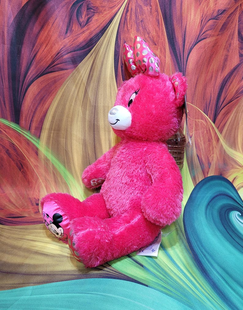 Puede incluir: Un oso de peluche rosa brillante con un lazo en la cabeza y un gr&aacute;fico de Minnie Mouse en el pie. El oso tiene un hocico blanco y una expresi&oacute;n sonriente. El fondo es un dise&ntilde;o abstracto colorido.