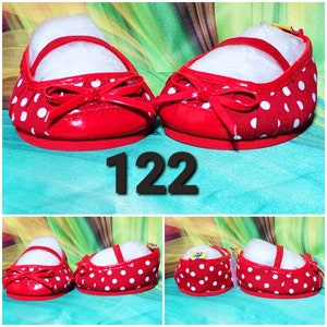K&ouml;nnte beinhalten: Ein Paar rote Puppenschuhe mit wei&szlig;en Punkten. Die Schuhe haben eine rote Schleife vorne und einen Riemen &uuml;ber dem Oberteil. Die Nummer "122" ist in Schwarz gedruckt.