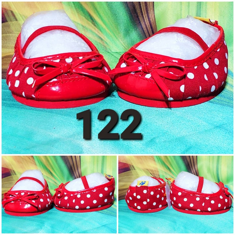 K&ouml;nnte beinhalten: Ein Paar rote Puppenschuhe mit wei&szlig;en Punkten. Die Schuhe haben eine rote Schleife vorne und einen Riemen &uuml;ber dem Oberteil. Die Nummer "122" ist in Schwarz gedruckt.